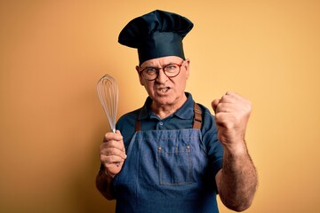 Fototapeta premium Middle age hoary cooker man wearing apron and hat holding whisk over yellow background annoyed and frustrated shouting with anger, crazy and yelling with raised hand, anger concept