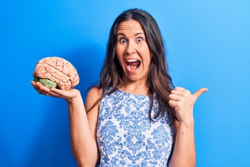 Young beautiful brunette woman asking for care memory holding brain over blue background pointing...
