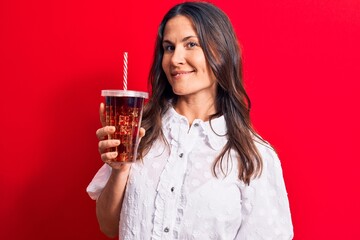 Beautiful brunette woman drinking cola refreshment beverage using straw over red background looking positive and happy standing and smiling with a confident smile showing teeth