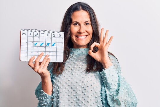 Young beautiful brunette woman holding travel calendar reminder over white background doing ok sign with fingers, smiling friendly gesturing excellent symbol