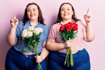 Young plus size twins holding flowers surprised with an idea or question pointing finger with happy face, number one