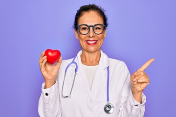 Middle age senior cardiologist doctor woman holding red heart over purple background very happy pointing with hand and finger to the side