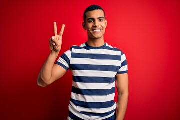 Fototapeta premium Handsome african american man wearing casual striped t-shirt standing over red background smiling looking to the camera showing fingers doing victory sign. Number two.