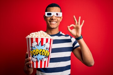 Young handsome african american man watching movie using 3d glasses eating popcorns smiling positive doing ok sign with hand and fingers. Successful expression.