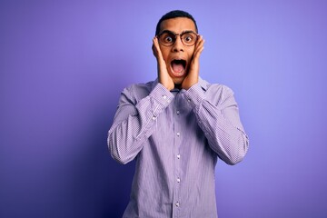 Handsome african american man wearing striped shirt and glasses over purple background afraid and shocked, surprise and amazed expression with hands on face