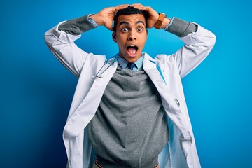 Handsome african american doctor man wearing coat and stethoscope over blue background Crazy and scared with hands on head, afraid and surprised of shock with open mouth