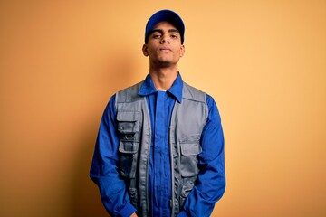 Young handsome african american handyman wearing worker uniform and cap with serious expression on face. Simple and natural looking at the camera.