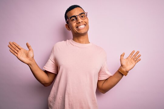 Handsome african american man wearing casual t-shirt and glasses over pink background clueless and confused expression with arms and hands raised. Doubt concept.