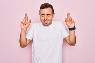 Fototapeta premium Handsome man with blue eyes wearing casual white t-shirt standing over pink background gesturing finger crossed smiling with hope and eyes closed. Luck and superstitious concept.