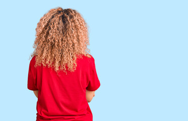 Young blonde woman with curly hair wearing casual red tshirt standing backwards looking away with...