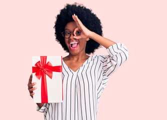 Young african american woman holding gift smiling happy doing ok sign with hand on eye looking through fingers