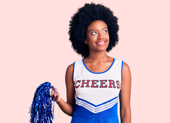 Young african american woman wearing cheerleader uniform holding pompom smiling looking to the side...