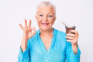 Senior beautiful woman with blue eyes and grey hair drinking mate infusion doing ok sign with fingers, smiling friendly gesturing excellent symbol