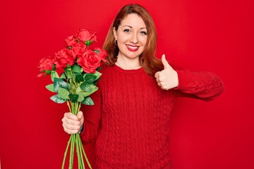 Young beautiful redhead woman holding bouquet of red roses flowers over isolated background happy with big smile doing ok sign, thumb up with fingers, excellent sign