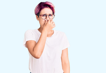 Young beautiful woman with pink hair wearing casual clothes and glasses smelling something stinky and disgusting, intolerable smell, holding breath with fingers on nose. bad smell