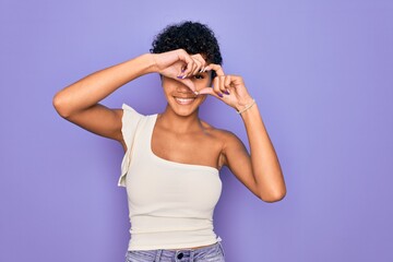 Young beautiful african american afro woman wearing casual t-shirt over purple background Doing heart shape with hand and fingers smiling looking through sign