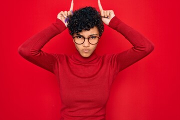 Fototapeta na wymiar Young beautiful african american afro woman wearing turtleneck sweater and glasses doing funny gesture with finger over head as bull horns