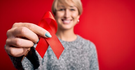 Young blonde woman with short hair holding hiv and aids awareness red ribbon with a happy face standing and smiling with a confident smile showing teeth - obrazy, fototapety, plakaty