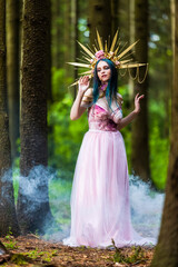 Obraz na płótnie Canvas Costume Play Concepts. Sexy and Sensual Crowned Forest Nymph with Flowery Golden Crown Posing in Smoky Summer Forest Against High Trees.
