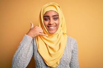 Young beautiful african american girl wearing muslim hijab over isolated yellow background smiling doing phone gesture with hand and fingers like talking on the telephone. Communicating concepts.