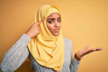 Young beautiful african american girl wearing muslim hijab over isolated yellow background confused and annoyed with open palm showing copy space and pointing finger to forehead. Think about it.