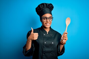 Fototapeta premium Young african american chef girl wearing cooker uniform and hat holding wooden spoon tool happy with big smile doing ok sign, thumb up with fingers, excellent sign