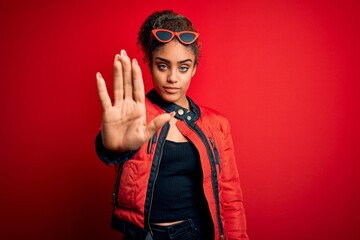 Obraz premium Beautiful african american girl wearing red jacket and sunglasses over isolated background doing stop sing with palm of the hand. Warning expression with negative and serious gesture on the face.