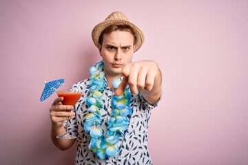 Young redhead tourist man on vacation wearing hat and hawaiian lei drinking cocktail pointing with...