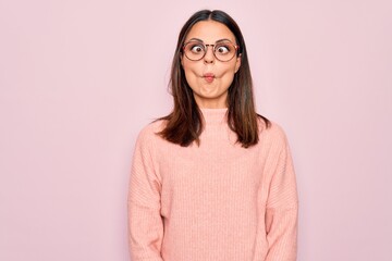 Young beautiful brunette woman wearing casual sweater and glasses over pink background making fish...