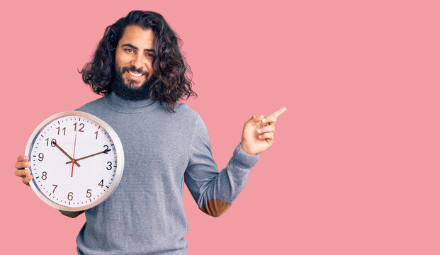 Young arab man holding big clock smiling happy pointing with hand and finger to the side