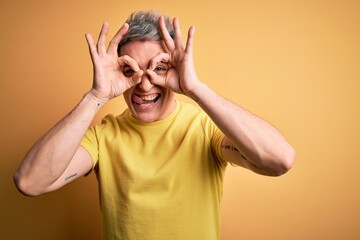 Young handsome modern man wearing yellow shirt over yellow isolated background doing ok gesture like binoculars sticking tongue out, eyes looking through fingers. Crazy expression.