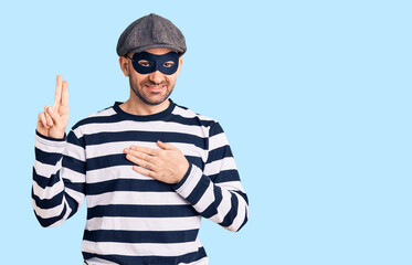 Young handsome man wearing burglar mask smiling swearing with hand on chest and fingers up, making a loyalty promise oath