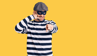 Young handsome man wearing burglar mask smiling doing talking on the telephone gesture and pointing to you. call me.