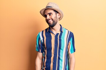 Young handsome man with beard wearing summer hat and shirt looking away to side with smile on face, natural expression. laughing confident.