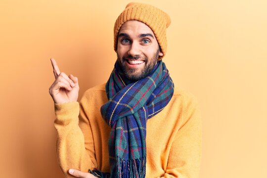 Young handsome man with beard wearing wool cap and scarf smiling happy pointing with hand and finger to the side