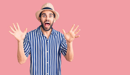 Naklejka premium Young handsome man with beard wearing summer hat and striped shirt celebrating crazy and amazed for success with arms raised and open eyes screaming excited. winner concept