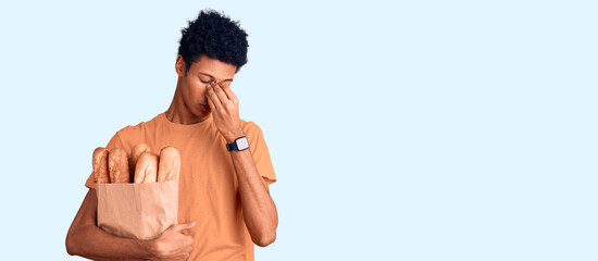 Young african american man holding paper bag with bread tired rubbing nose and eyes feeling fatigue and headache. stress and frustration concept.