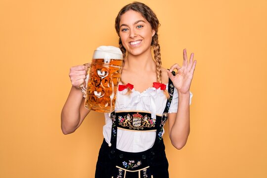 Beautiful blonde german woman with blue eyes wearing octoberfest dress drinking jar of beer doing ok sign with fingers, excellent symbol