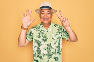 Middle age senior grey-haired man wearing summer hat and floral shirt on beach vacation showing and pointing up with fingers number seven while smiling confident and happy.