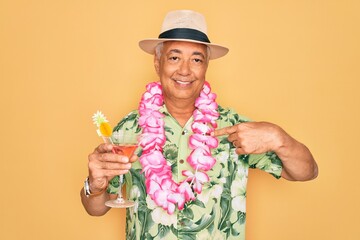 Middle age senior grey-haired man wearing summer hat and hawaiian lei drinking a cocktail with...