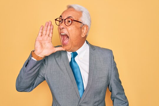 Middle age senior grey-haired handsome business man wearing glasses over yellow background shouting and screaming loud to side with hand on mouth. Communication concept.