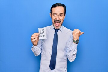 Middle age business man asking for social rights holding paper with we are equal message pointing thumb up to the side smiling happy with open mouth