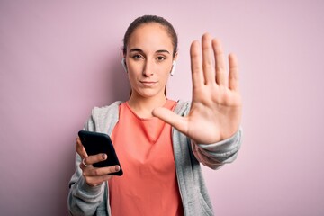Fototapeta premium Beautiful sporty woman doing sport listening to music using earphones over pink background with open hand doing stop sign with serious and confident expression, defense gesture