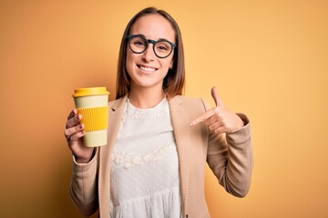 Young beautiful businesswoman drinking cup of takeaway coffee over yellow background with surprise face pointing finger to himself