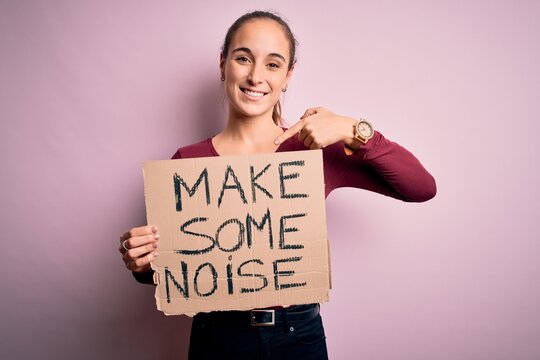 Beautiful activist woman holding banner with make some noise message over pink background with surprise face pointing finger to himself