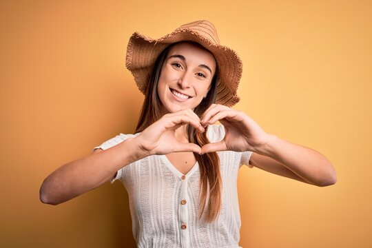 Young beautiful woman wearing casual t-shirt and summer hat over isolated yellow background smiling in love showing heart symbol and shape with hands. Romantic concept.
