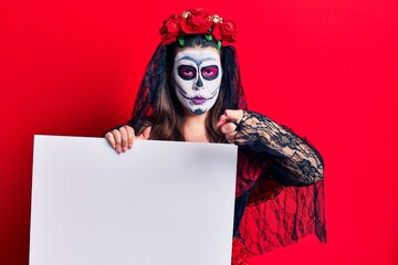 Young woman wearing day of the dead costume holding blank empty banner pointing with finger to the camera and to you, confident gesture looking serious