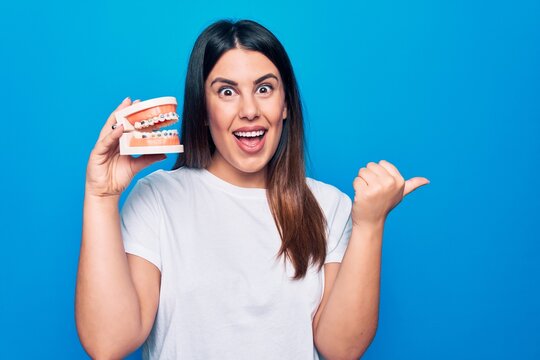 Young beautiful brunette woman asking for dental care holding denture with backets pointing thumb up to the side smiling happy with open mouth