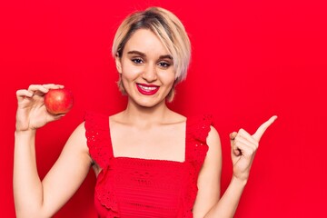 Young beautiful blonde woman holding red apple smiling happy pointing with hand and finger to the side