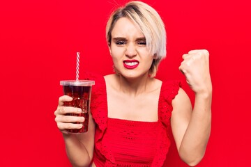 Young beautiful blonde woman drinking glass of cola beverage annoyed and frustrated shouting with anger, yelling crazy with anger and hand raised